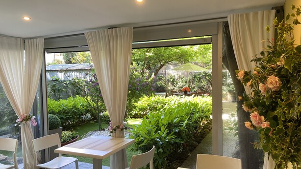 A dining area with a table and chairs, a view of a garden with plants and a table with flowers, and a curtain partially covering a window. - Paradise Inn (Livorno)
