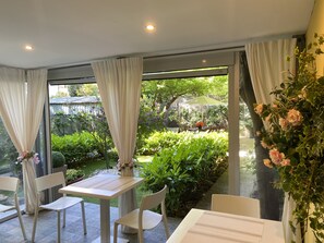 A dining area with a table and chairs, a view of a garden with plants and a table with flowers, and a curtain partially covering a window. - Paradise Inn (Livorno)