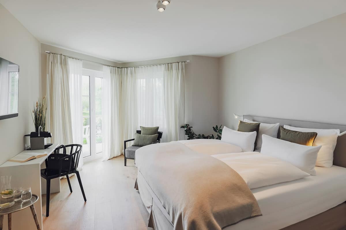 Ein modernes Hotelzimmer mit einem großen Bett, einem Schreibtisch mit Stuhl und Blick ins Freie durch große Fenster.