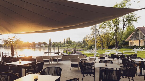 Un espace extérieur aménagé en terrasse, avec des tables et des chaises, offrant une vue sur un plan d’eau et une zone herbeuse dotée de chaises longues.