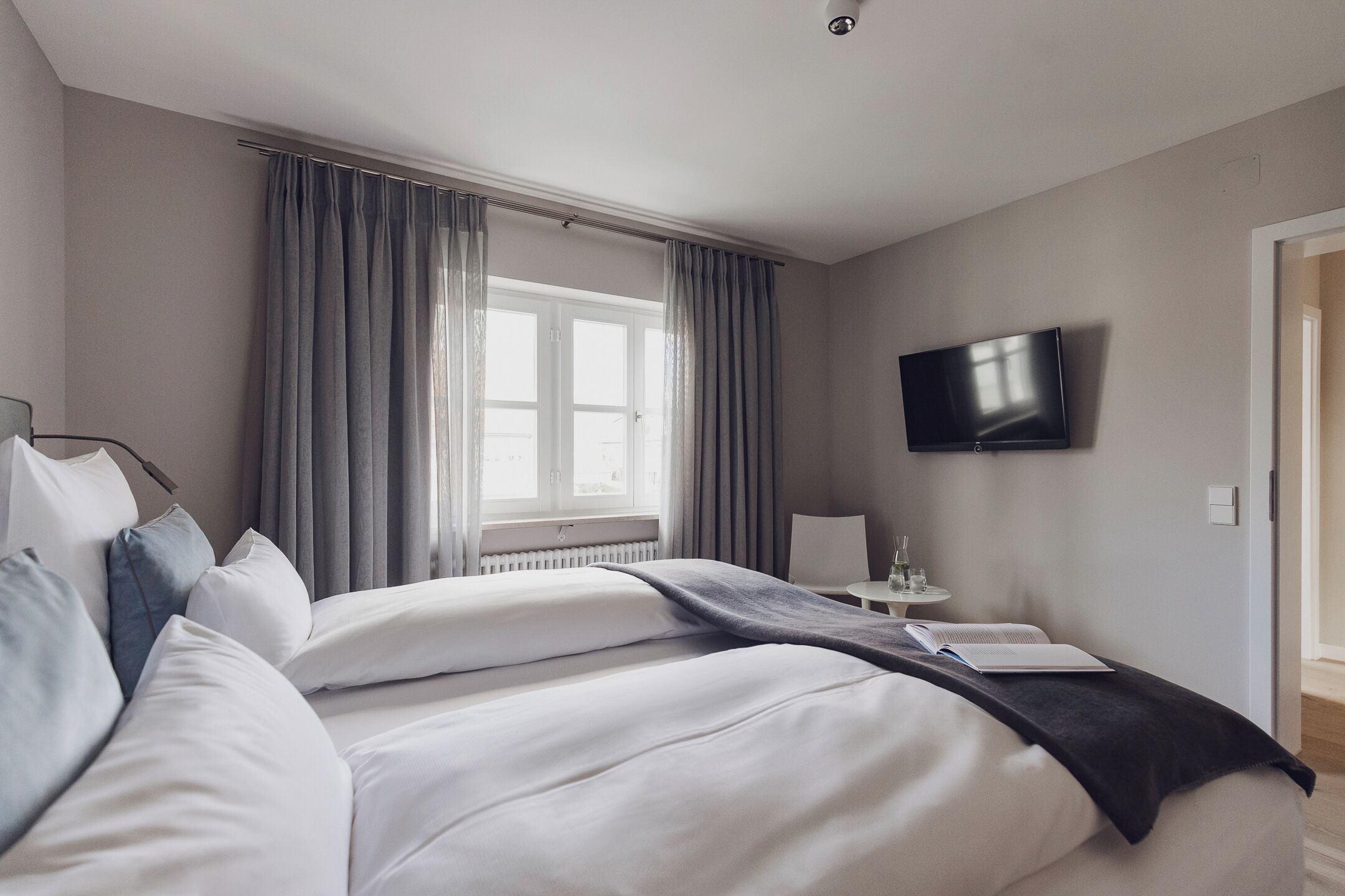 Una habitación de hotel moderna con una cama grande, un televisor de pantalla plana, una mesita con una silla y un ventanal con cortinas.