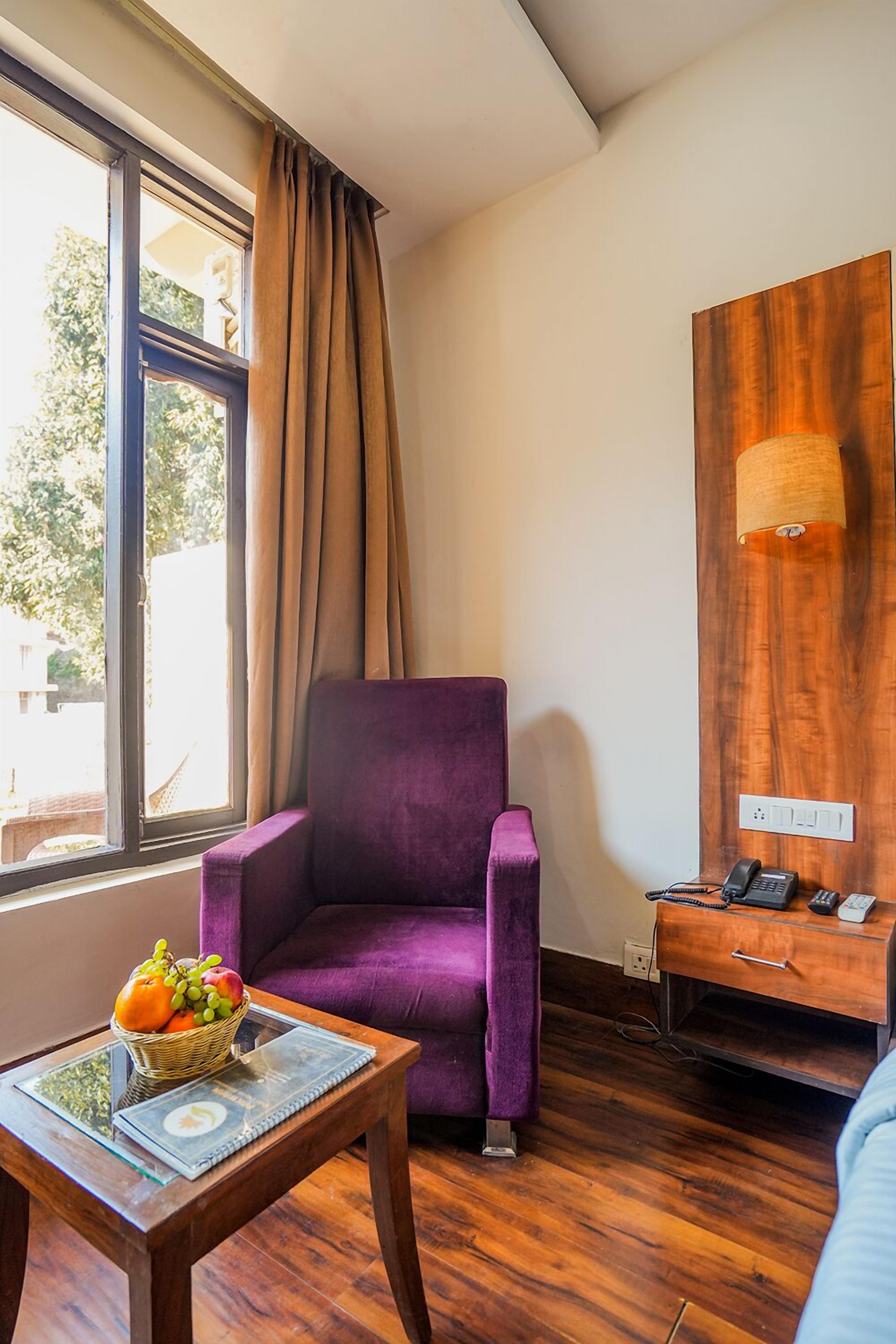 Une pièce avec un fauteuil violet, une table en bois sur laquelle se trouve un panier de fruits, un bureau en bois avec une lampe et une grande fenêtre avec des rideaux.