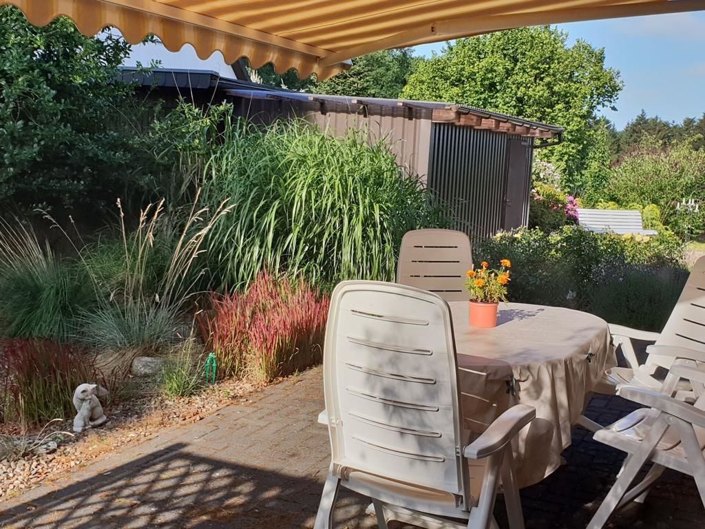Un patio exterior con mesa y sillas, rodeado de abundante follaje y una estructura de madera.