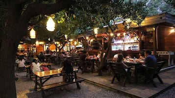 An outdoor dining area with wooden tables and benches, surrounded by trees and illuminated by hanging lights.