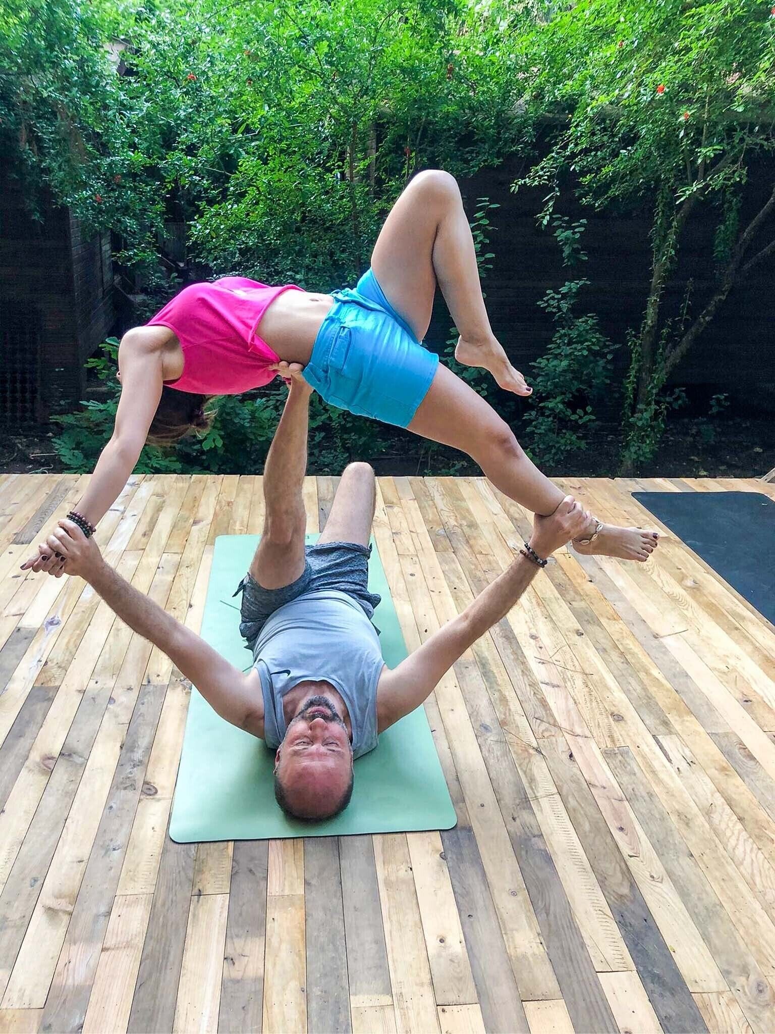 A man and a woman performing an acrobatic pose on a wooden deck with a green mat, surrounded by greenery.