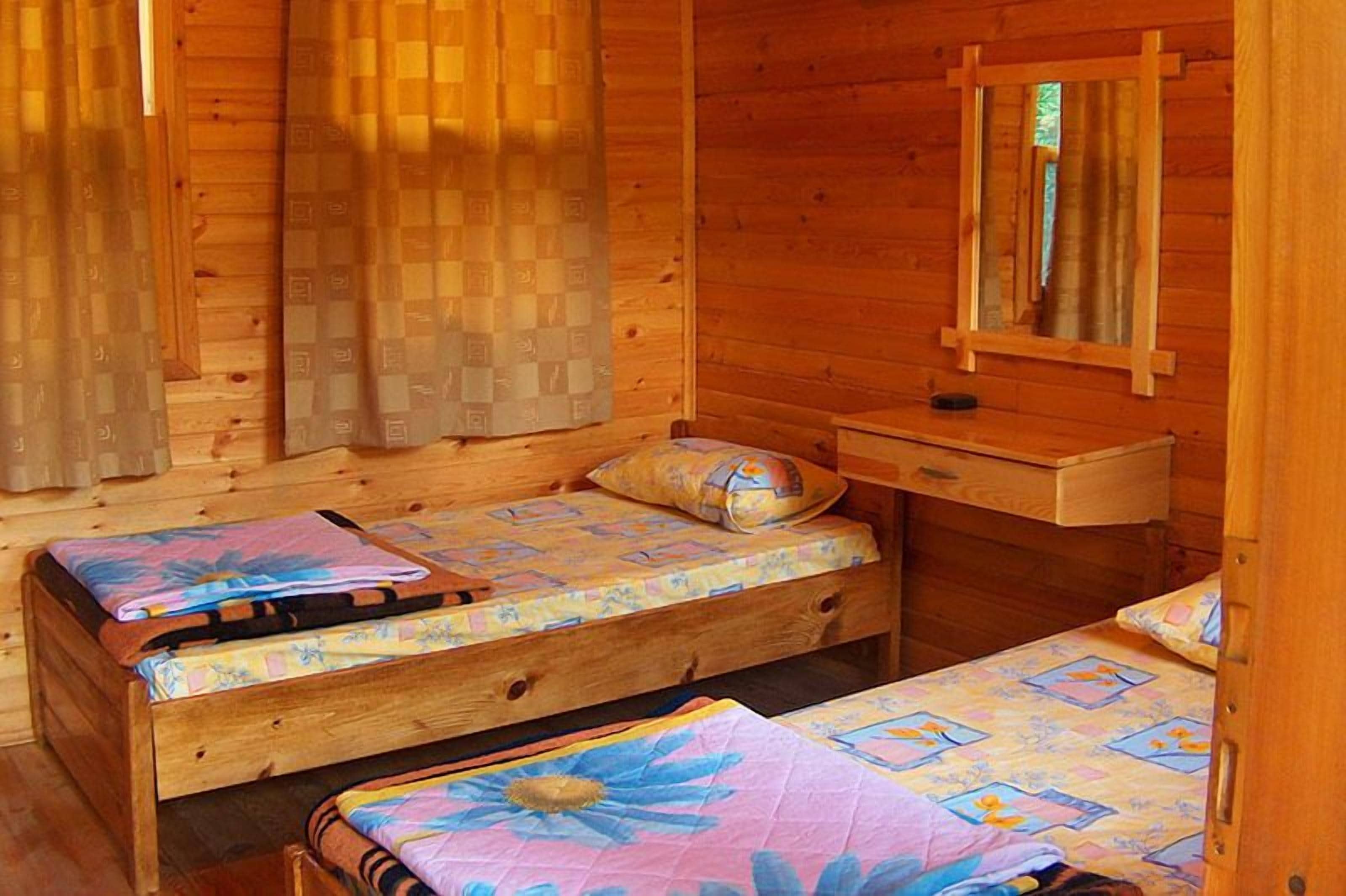Une chambre dans un chalet en bois, avec deux lits simples, une petite commode en bois, un miroir et une fenêtre avec des rideaux.
