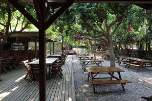 An outdoor restaurant with wooden tables and benches, surrounded by trees and covered by a wooden pergola. - Sheriff Olimpos (Kumluca)