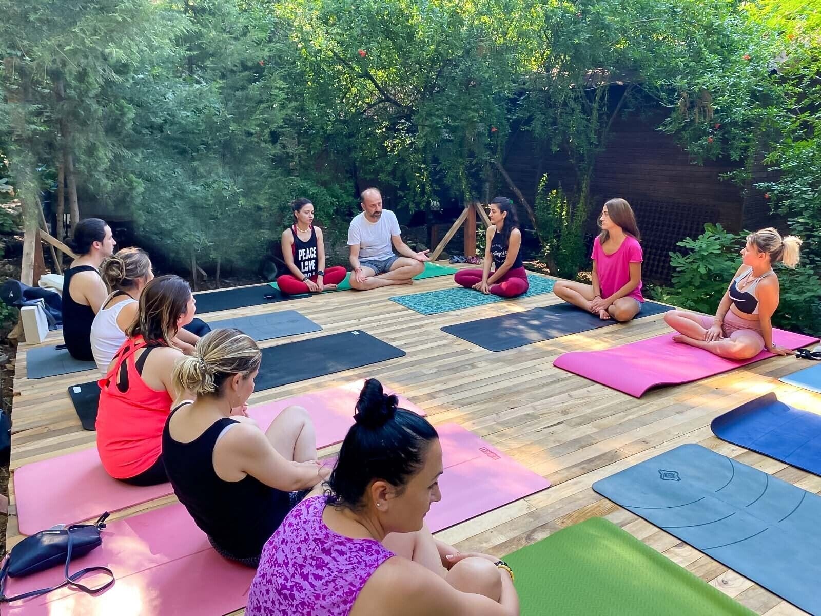 A group of people sitting on yoga mats in an outdoor setting with a wooden deck and greenery in the background.
