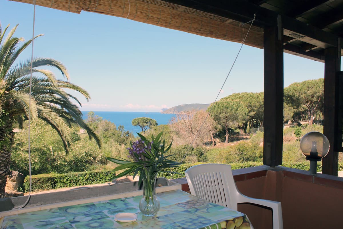 A table with a vase of flowers, a chair, and a view of the sea and hills.