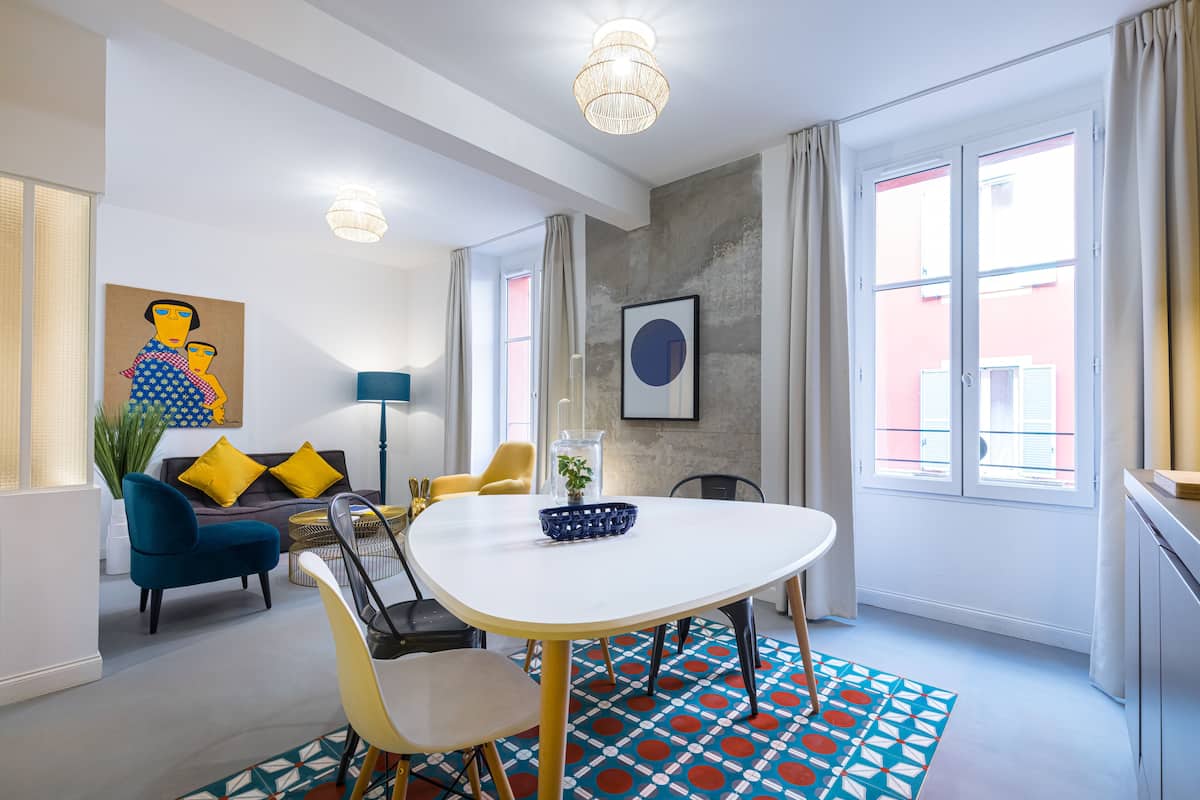 A modern living room with a white table, blue and yellow chairs, a sofa with yellow cushions, and a large window with curtains.
