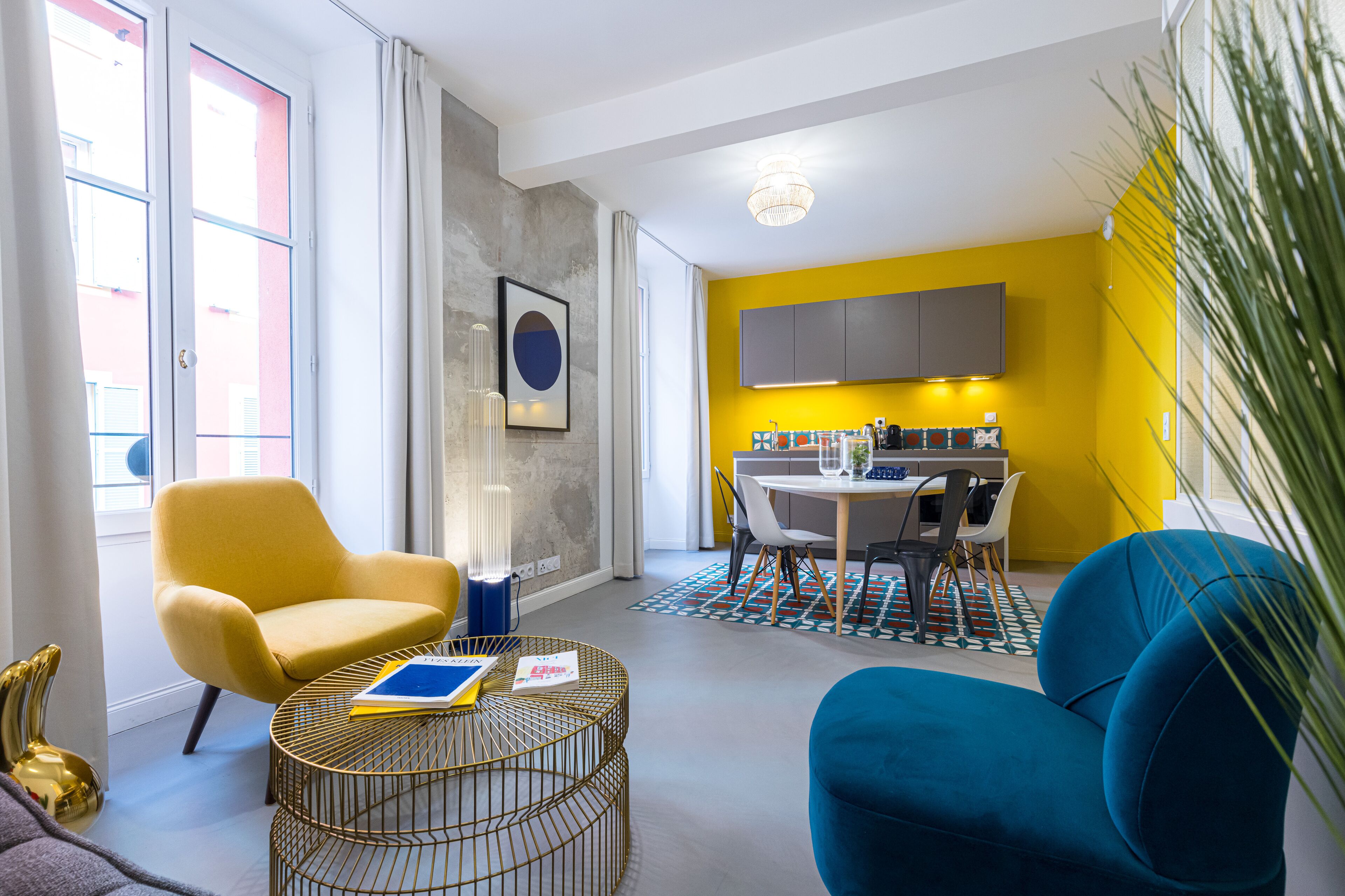 A modern living room with a yellow chair, a blue sofa, and a round coffee table. There is a dining area with a table and chairs, and a kitchen with cabinets.