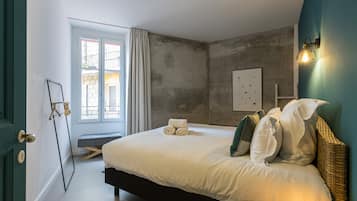Quarto com uma cama grande, um banquinho, uma luminária na parede e uma janela com cortinas.