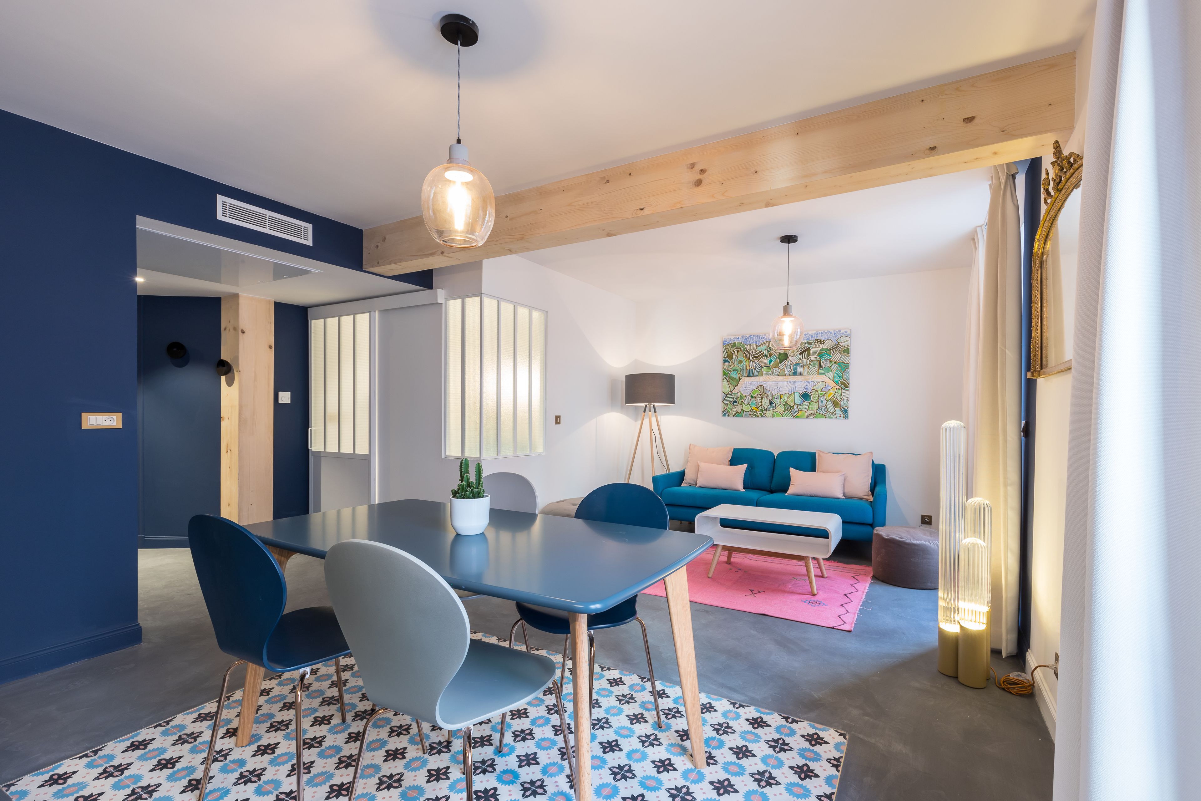 A modern living room with a blue sofa, a dining table with chairs, a floor lamp, and a painting on the wall.