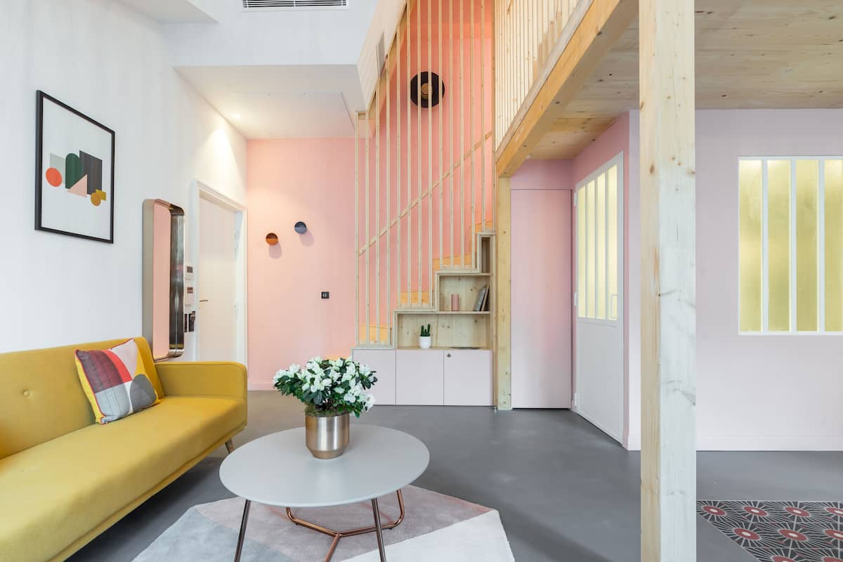 A modern living room with a yellow sofa, a round coffee table with a flower vase, and a staircase with a pink railing.