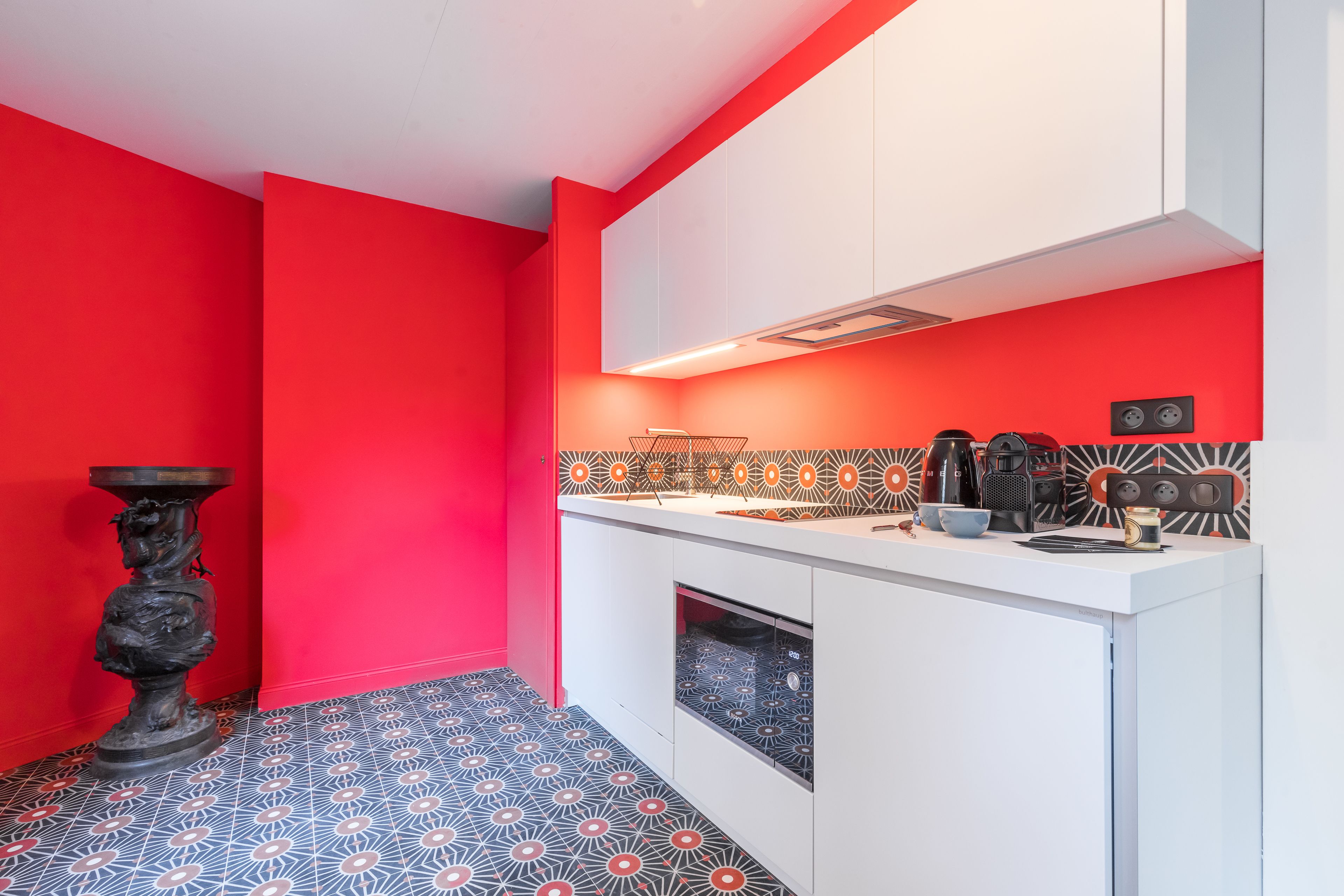 Una cucina compatta con mobili bianchi, una parete di colore rosso e piastrelle a pavimento decorate.