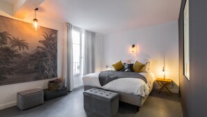 A bedroom with a large bed, bedside lamps, a grey bench, a wall-mounted shelf, and a large painting. - L'Abeille Boutique Apartments (Nice)