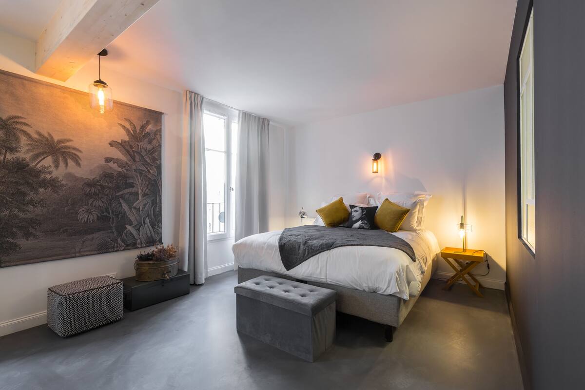 A bedroom with a large bed, bedside lamps, a grey bench, a wall-mounted shelf, and a large painting.