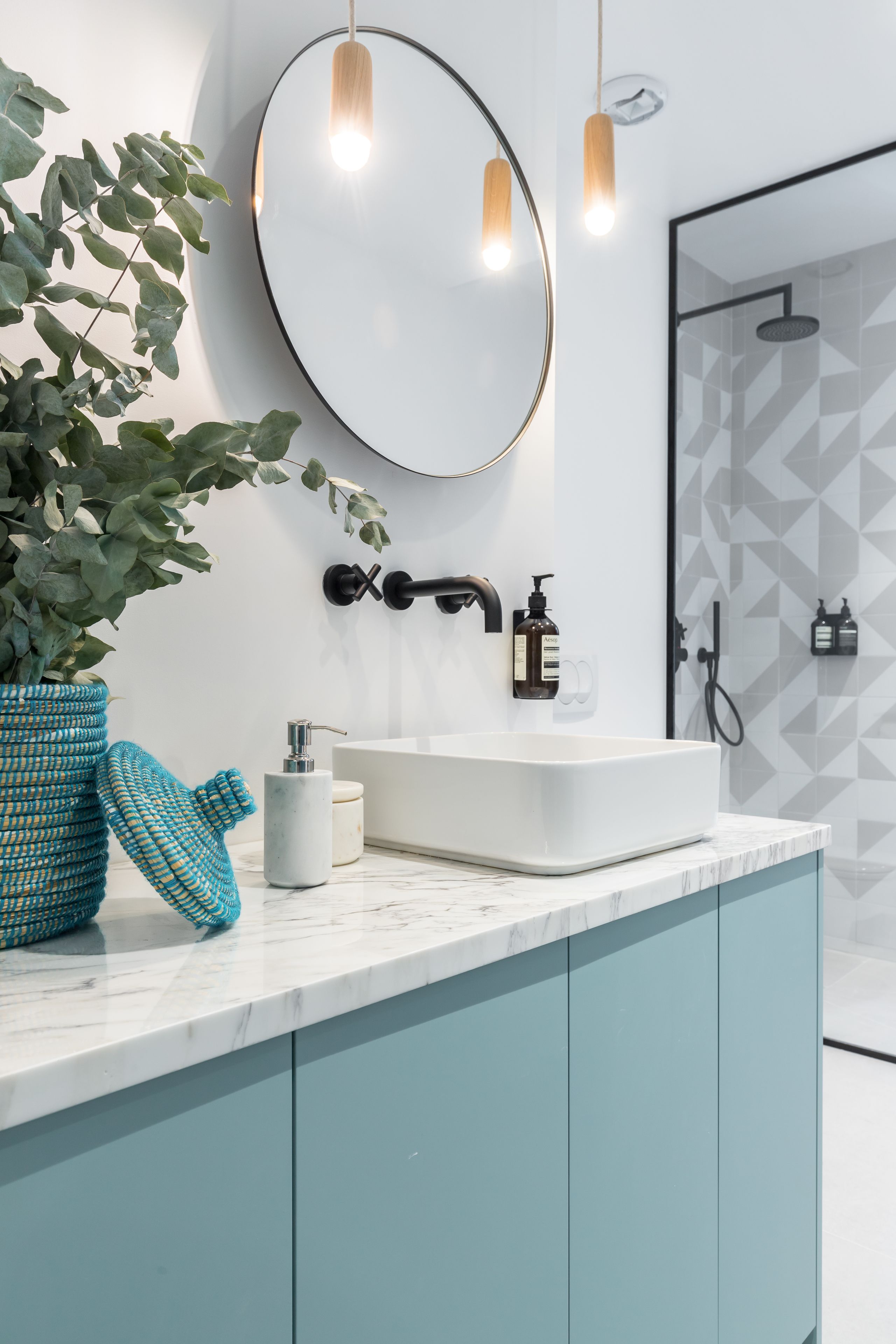 A modern bathroom with a white sink, teal cabinets, a round mirror, and a potted plant.