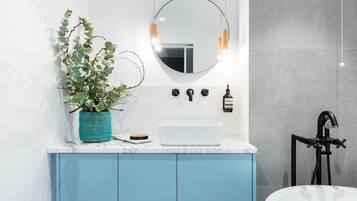 A modern bathroom with a white sink, blue cabinets, a round mirror with pendant lights, and a black handheld showerhead.