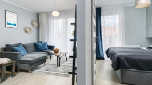 Ein modernes Hotelzimmer mit grauem Sofa, einem kleinen Tisch mit Obstschale, einem Bett mit dunkler Bettdecke und einem großen Fenster mit leichtem Vorhang.
