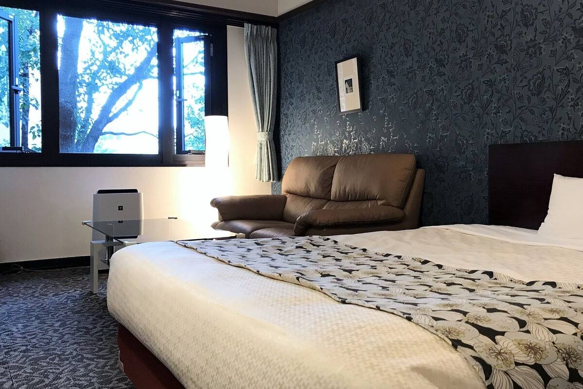 Ett hotellrum med en stor säng, en brun läderfåtölj, ett skrivbord med en skrivare och ett fönster med utsikt över träd.