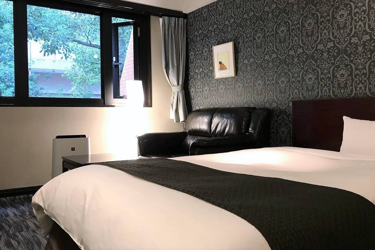 Ett hotellrum med en stor säng, en fåtölj i läder, ett nattduksbord och ett fönster med gardiner.