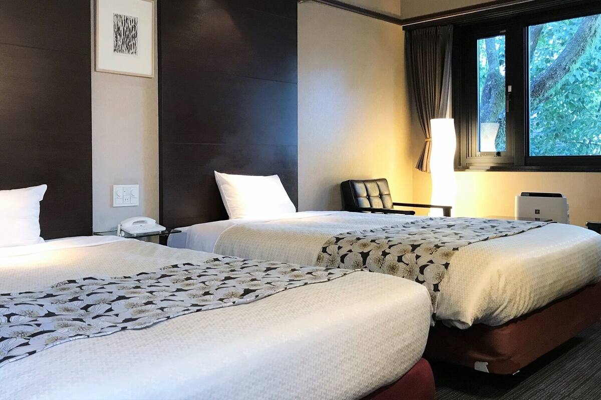 Ett hotellrum med två sängar, ett skrivbord, en stol, en lampa och ett fönster med utsikt över träd.