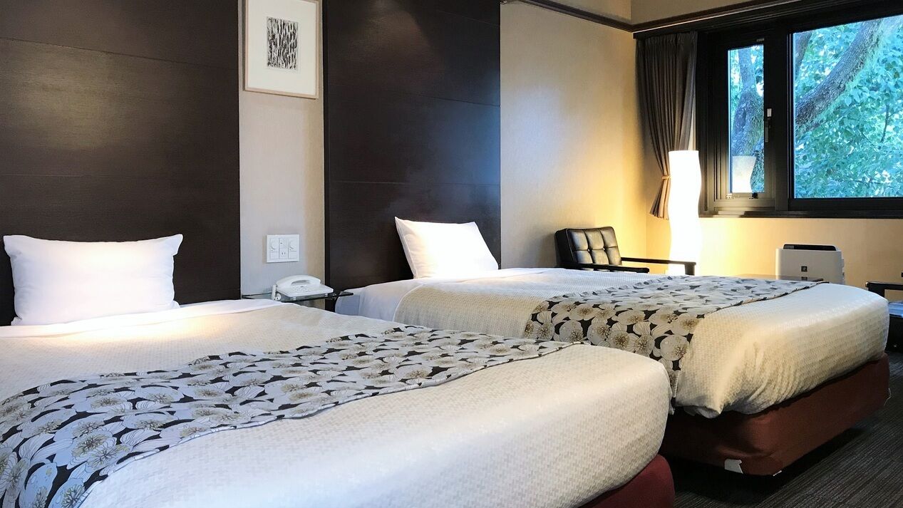 A hotel room with two beds, a desk, a chair, a lamp, and a window with a view of trees.