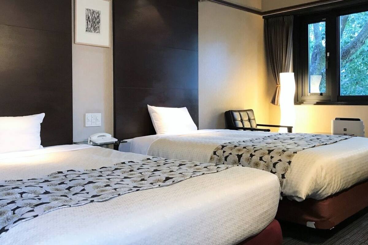 Ett hotellrum med två sängar, ett skrivbord, en stol, en lampa och ett fönster med utsikt över träd.