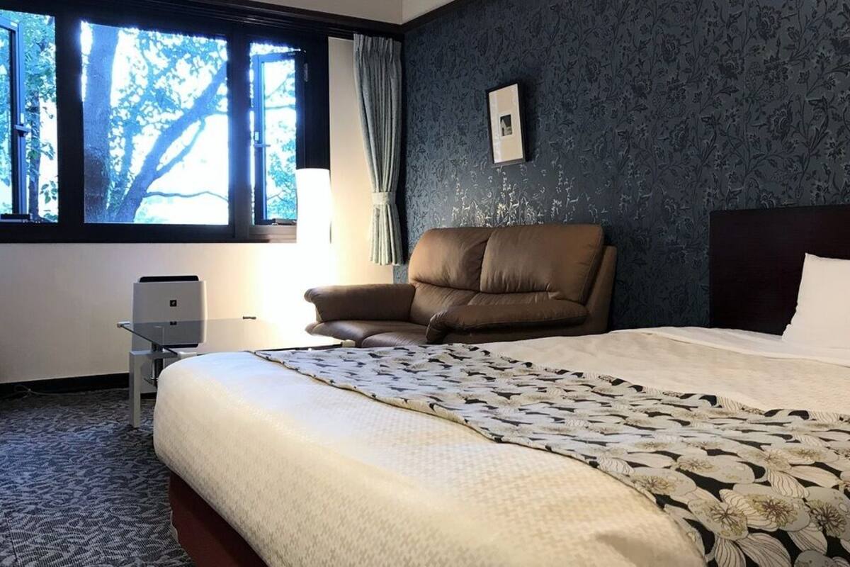 Ett hotellrum med en stor säng, en stol, ett skrivbord med en skrivare och ett fönster med gardiner.