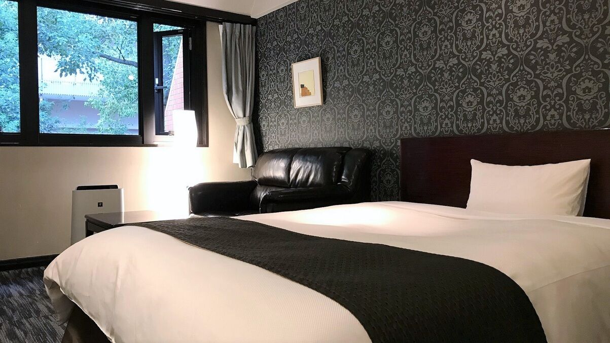 A hotel room with a large bed, a black leather chair, a nightstand, a lamp, and a window with curtains.