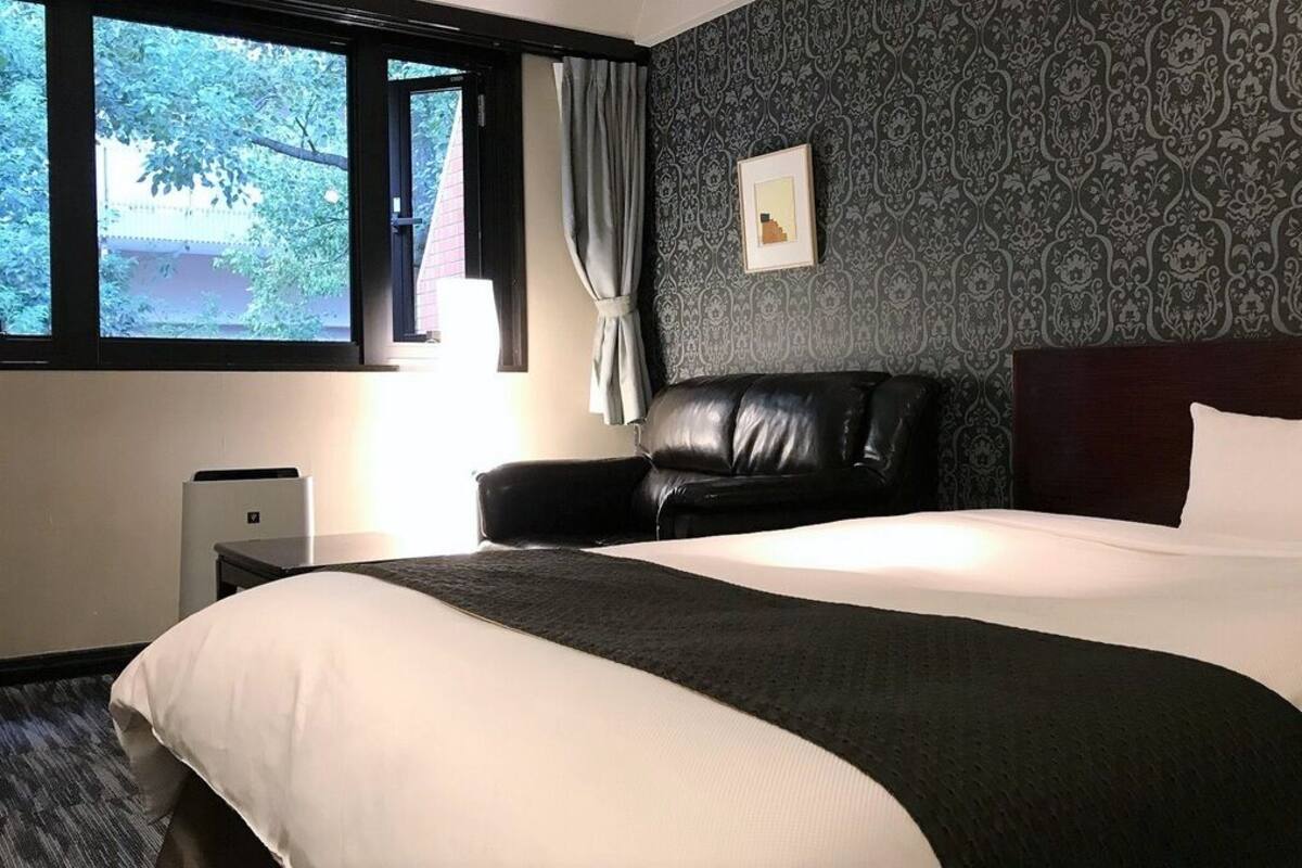 Ett hotellrum med en stor säng, en svart läderfåtölj, ett nattduksbord, en lampa och ett fönster med gardiner.