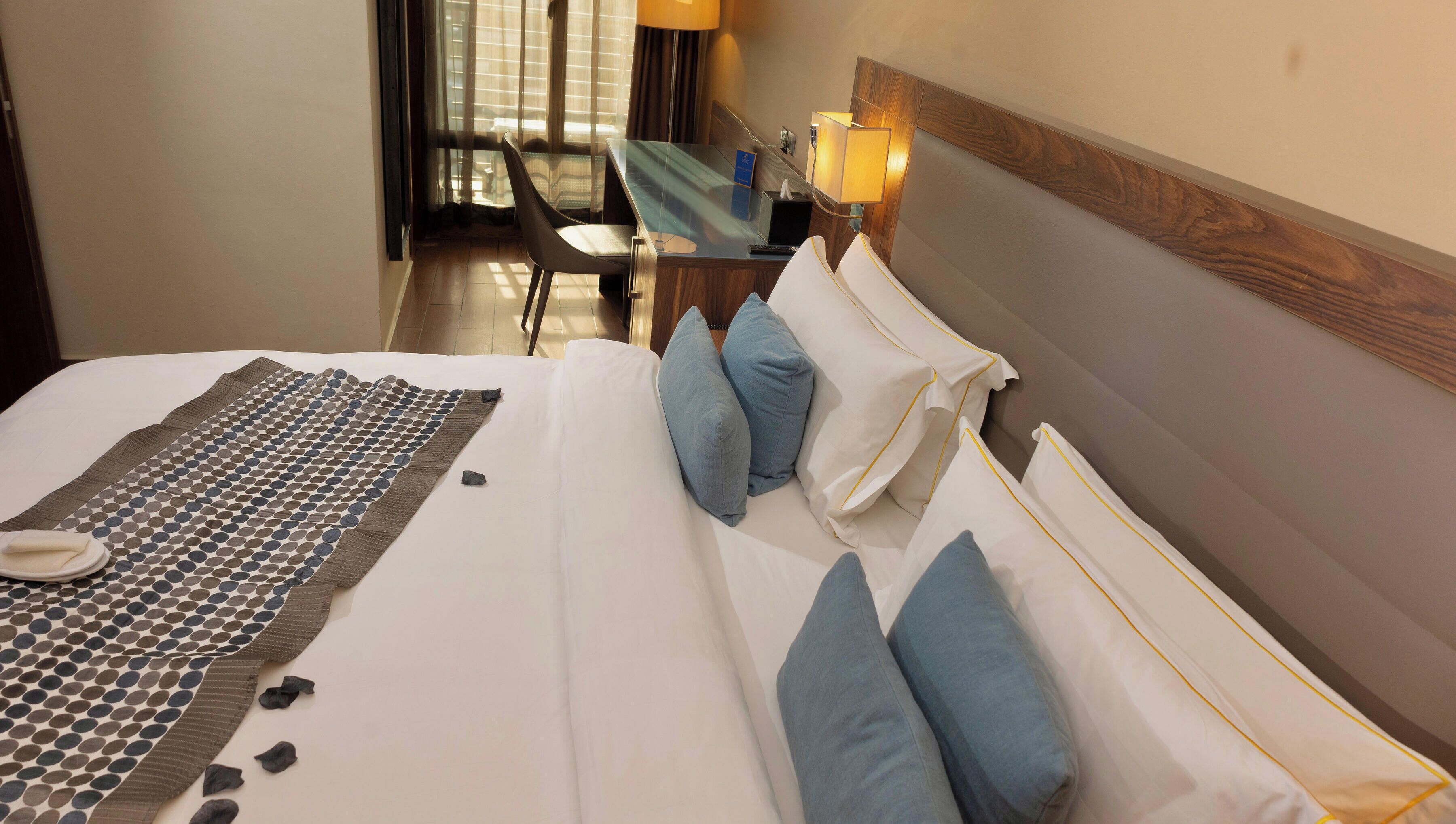 A hotel room with a bed, pillows, a bedside table, a lamp, and a view of the outside.