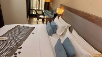 Habitación de hotel con cama, almohadas, mesita de noche, lámpara y vistas al exterior.