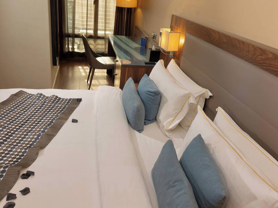 Habitación de hotel con cama, almohadas, mesita de noche, lámpara y vistas al exterior.