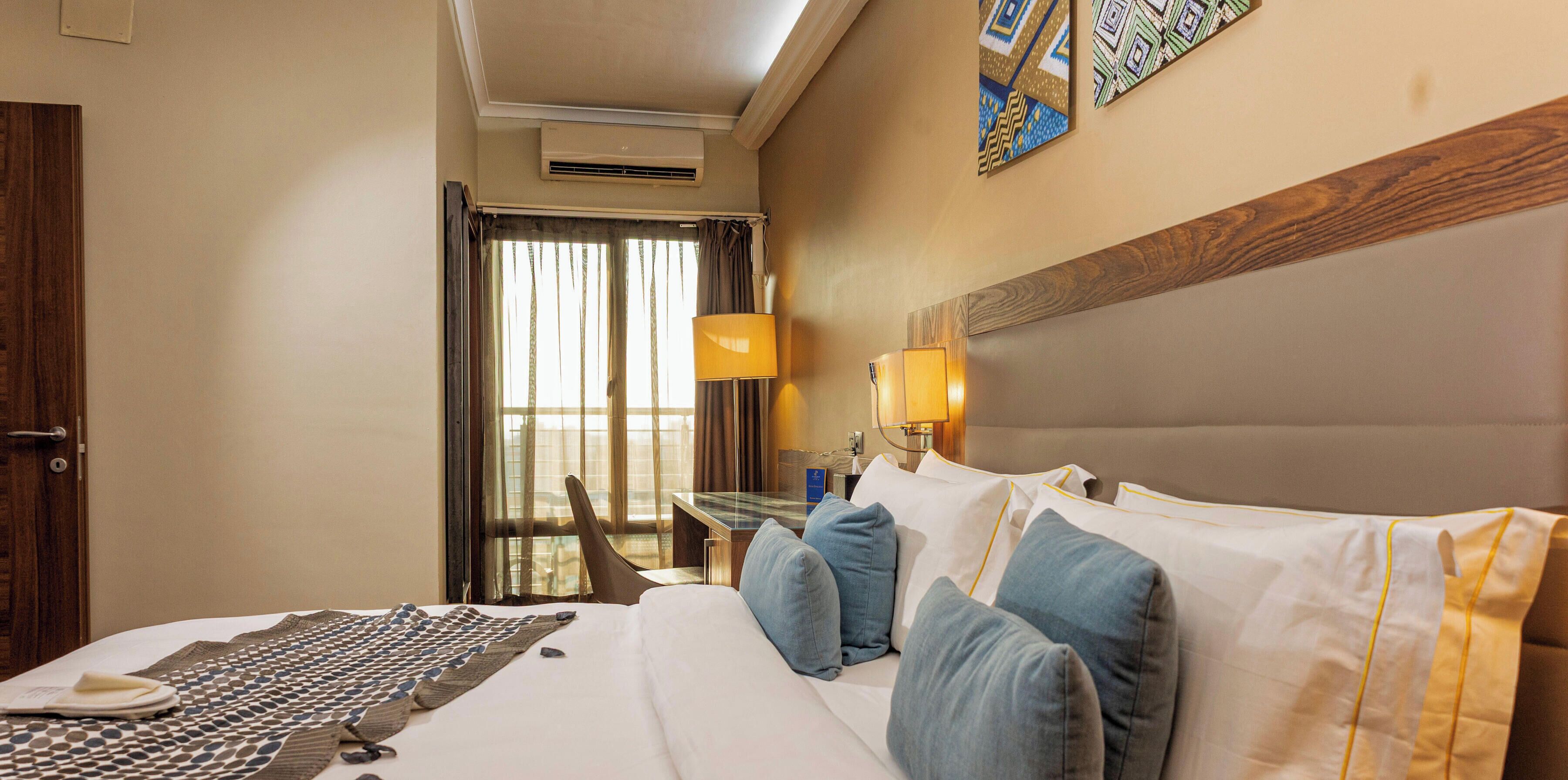 Una habitación de hotel con cama, escritorio, silla, lámpara y ventana con cortinas.
