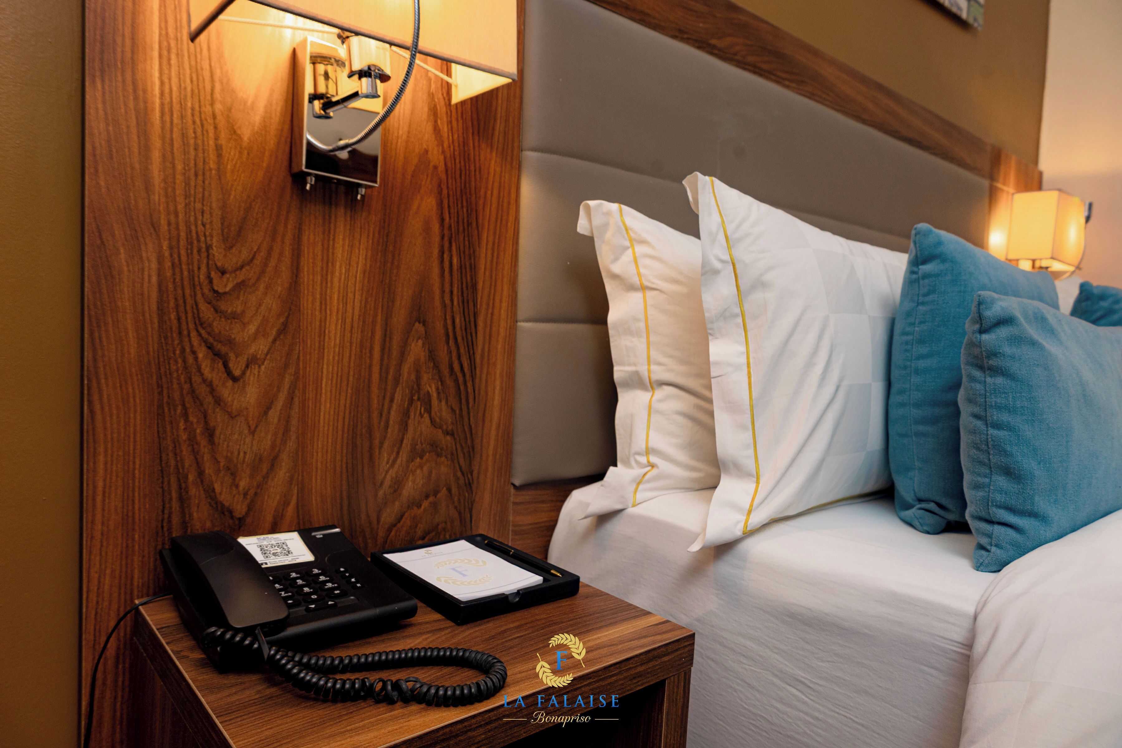 A hotel room with a wooden nightstand, a black telephone, a framed certificate, and a bed with white and blue pillows.