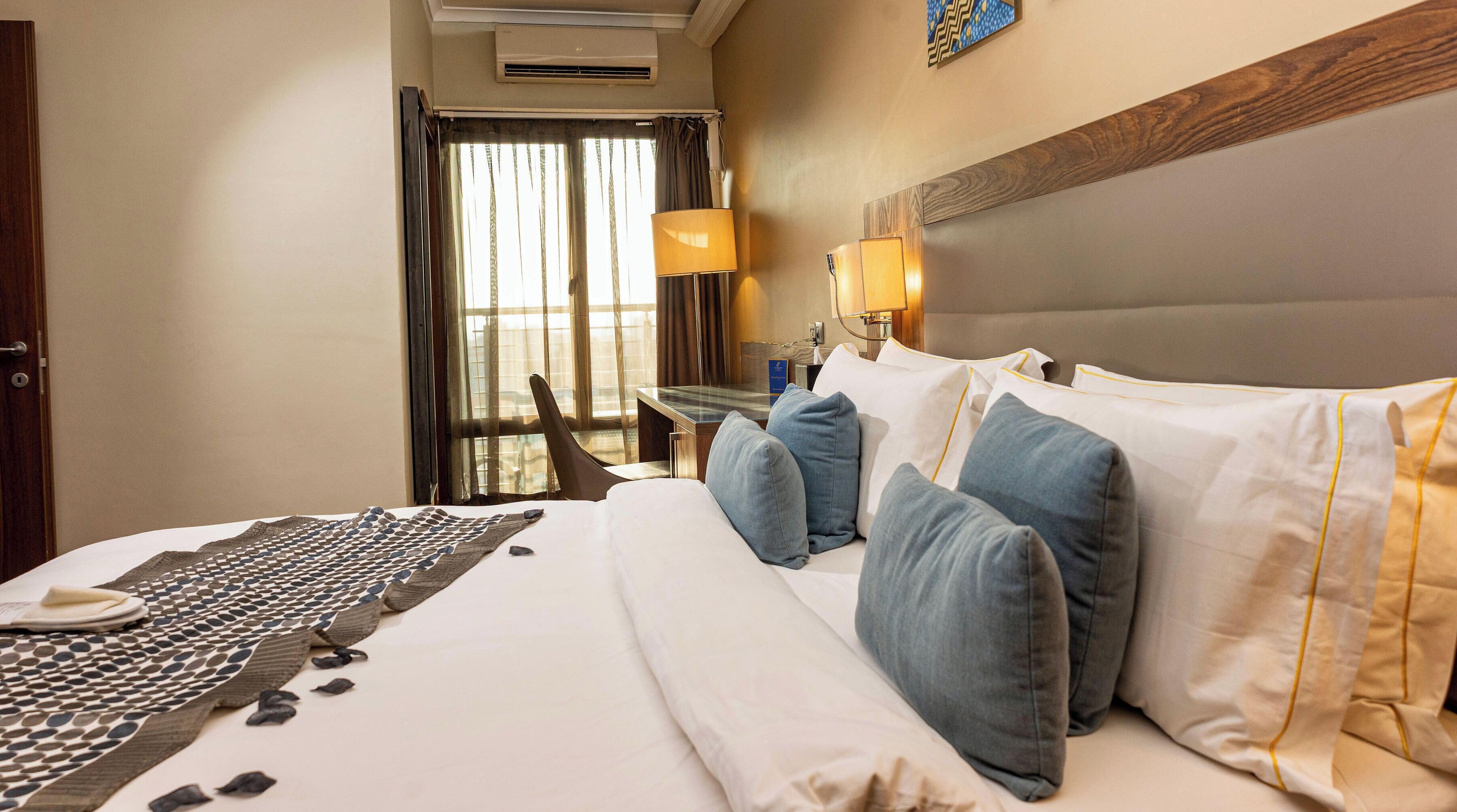 Habitación de hotel con cama, almohadas, mesita de noche, lámpara y vistas al exterior por la ventana.