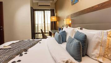 Habitación de hotel con cama, almohadas, mesita de noche, lámpara y vistas al exterior por la ventana.