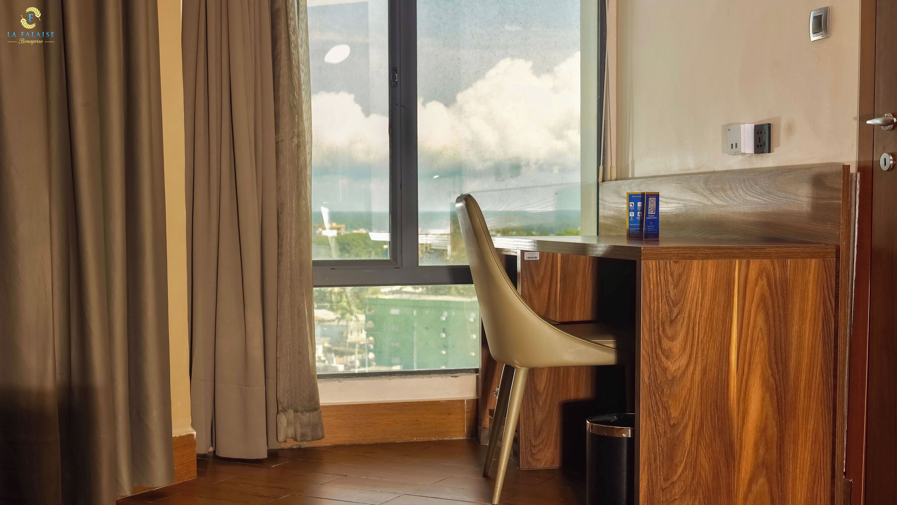 A hotel room with a large window, a wooden desk, a chair, and a trash bin.