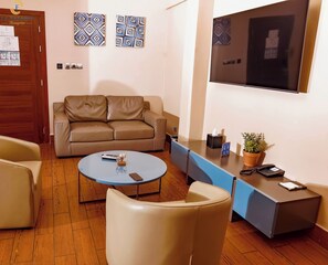 A living room with a sofa, a coffee table, a TV, and a desk with a phone and a plant. - Hotel La Falaise Bonapriso (Douala)