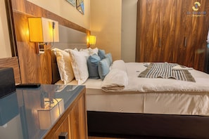 A hotel room with a bed, bedside lamps, a wooden wardrobe, and a desk with a remote control. - Hotel La Falaise Bonapriso (Douala)