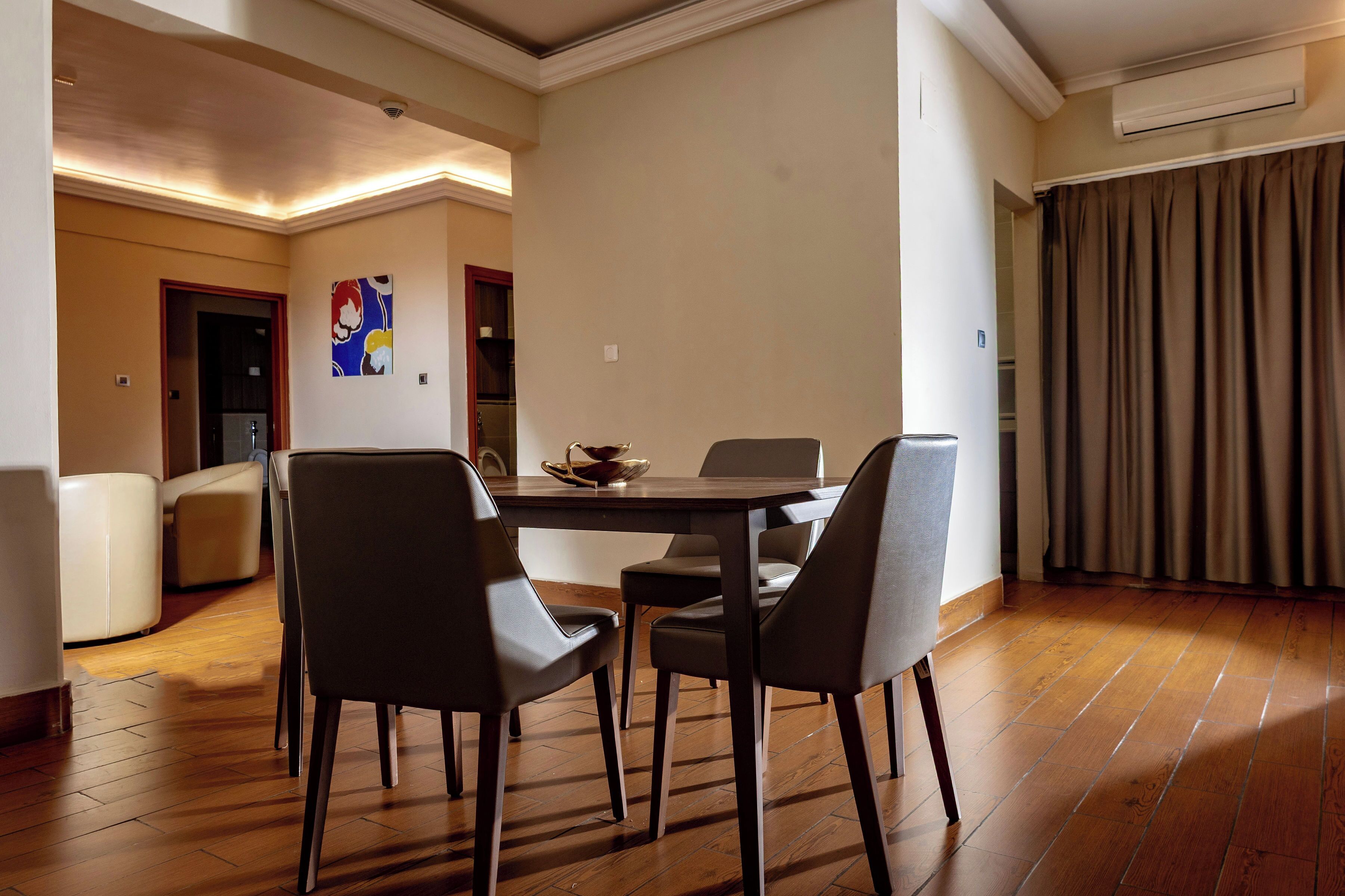 A dining area with a wooden table and four chairs, a painting on the wall, and an air conditioning unit.
