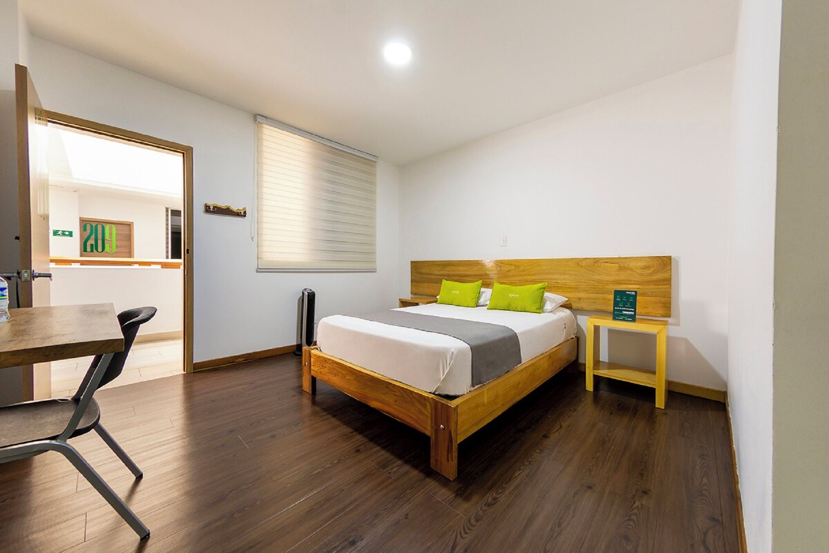 Ein Schlafzimmer mit einem hölzernen Bett, einem Nachttisch, einem kleinen Hocker, einem Schreibtisch mit Stuhl und einem Fenster mit Jalousien.