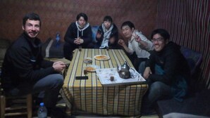 A group of people seated around a table with food and drinks, in a room with patterned wallpaper and a curtain. - Merzouga Experience Camp (Taouz)
