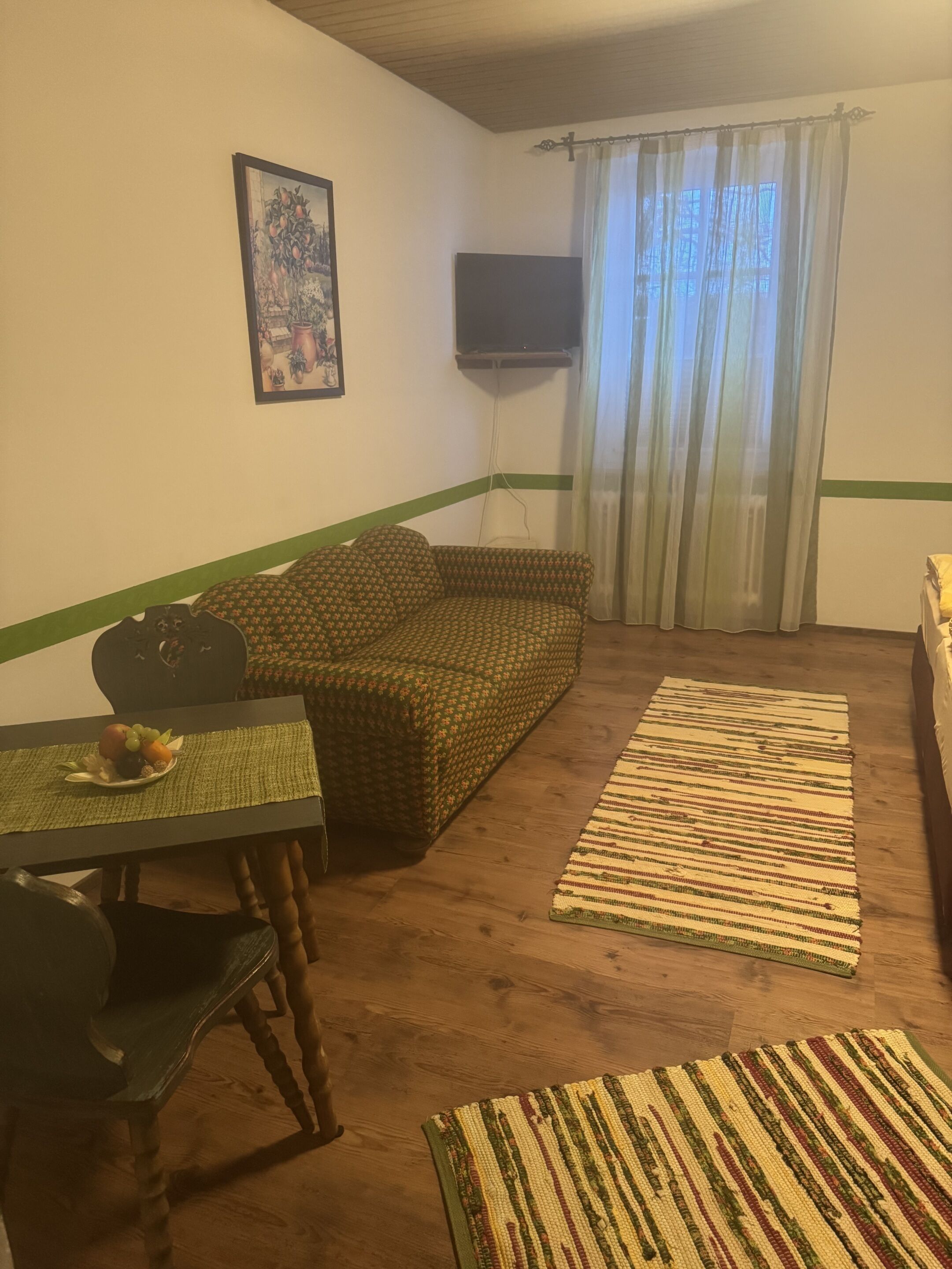 A cozy living room with a sofa, a television mounted on the wall, a dining table with a placemat, and a patterned rug on the floor.