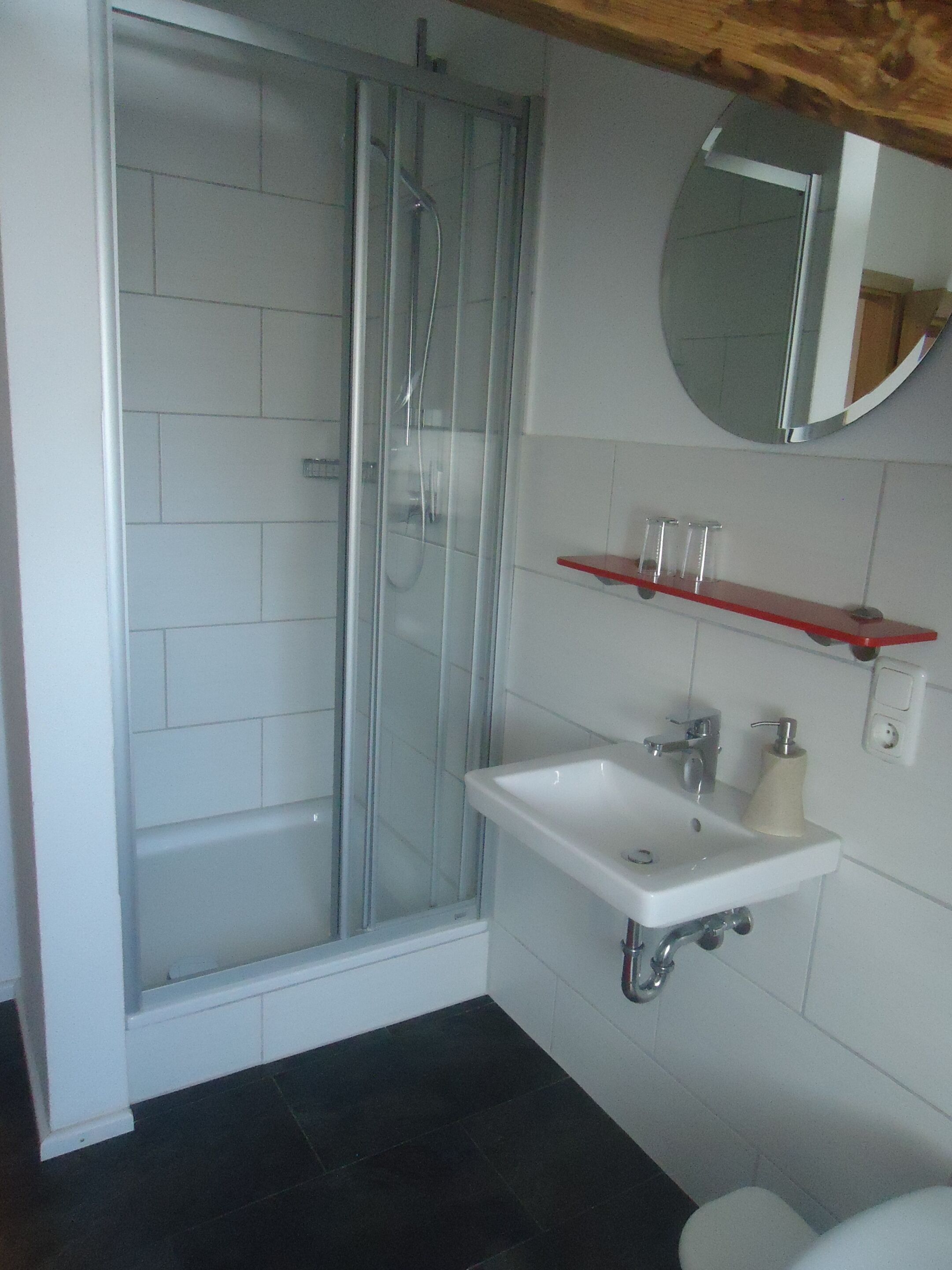 A bathroom with a glass-enclosed shower, a white sink with a mirror above it, and a red shelf.