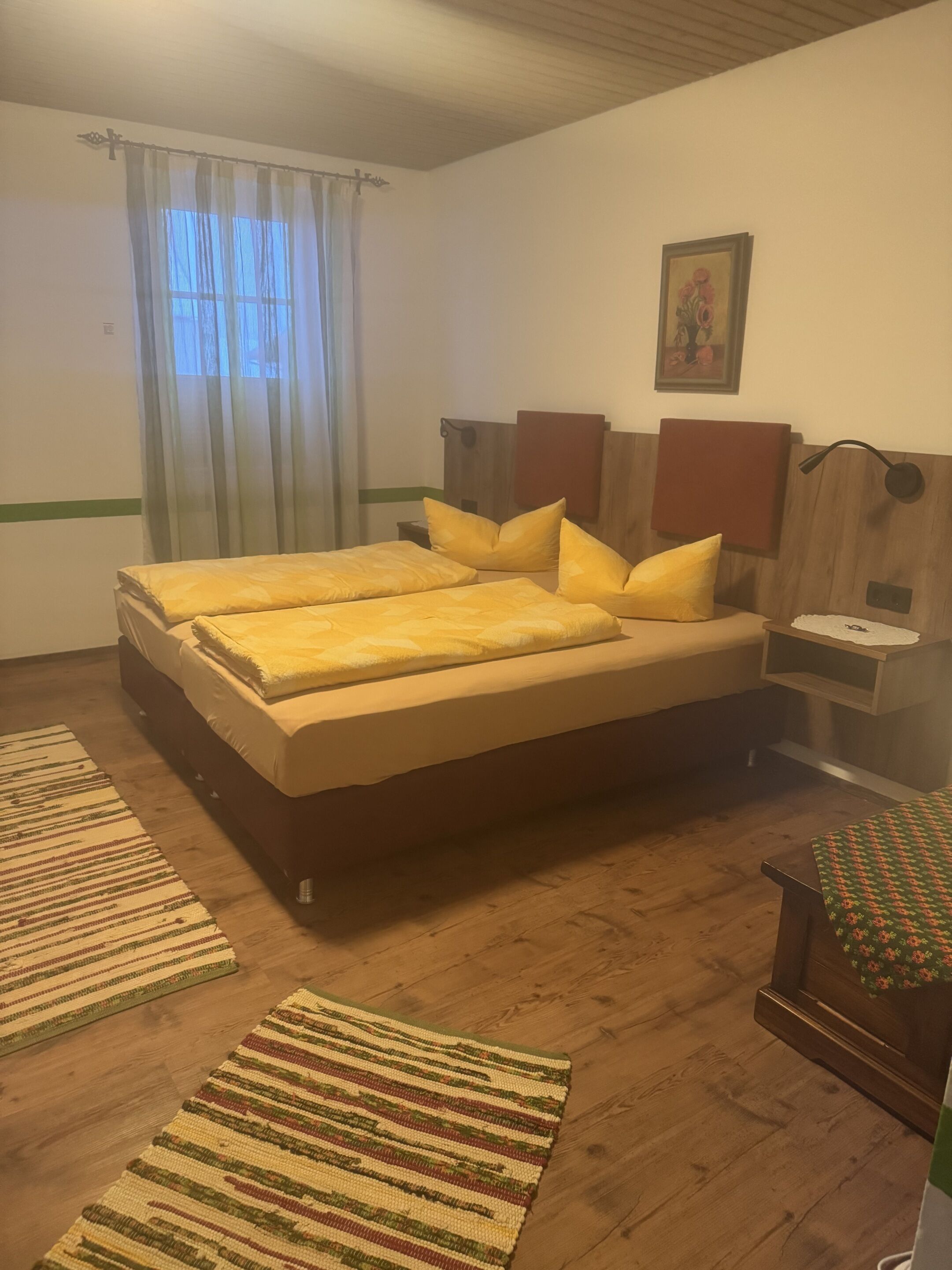 A double bed with yellow bedding, wooden headboard, two bedside tables with lamps, a painting on the wall, and a window with sheer curtains.
