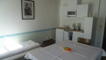 A small, simple room with a bed, a table with fruit, a kitchenette, and a framed picture on the wall.