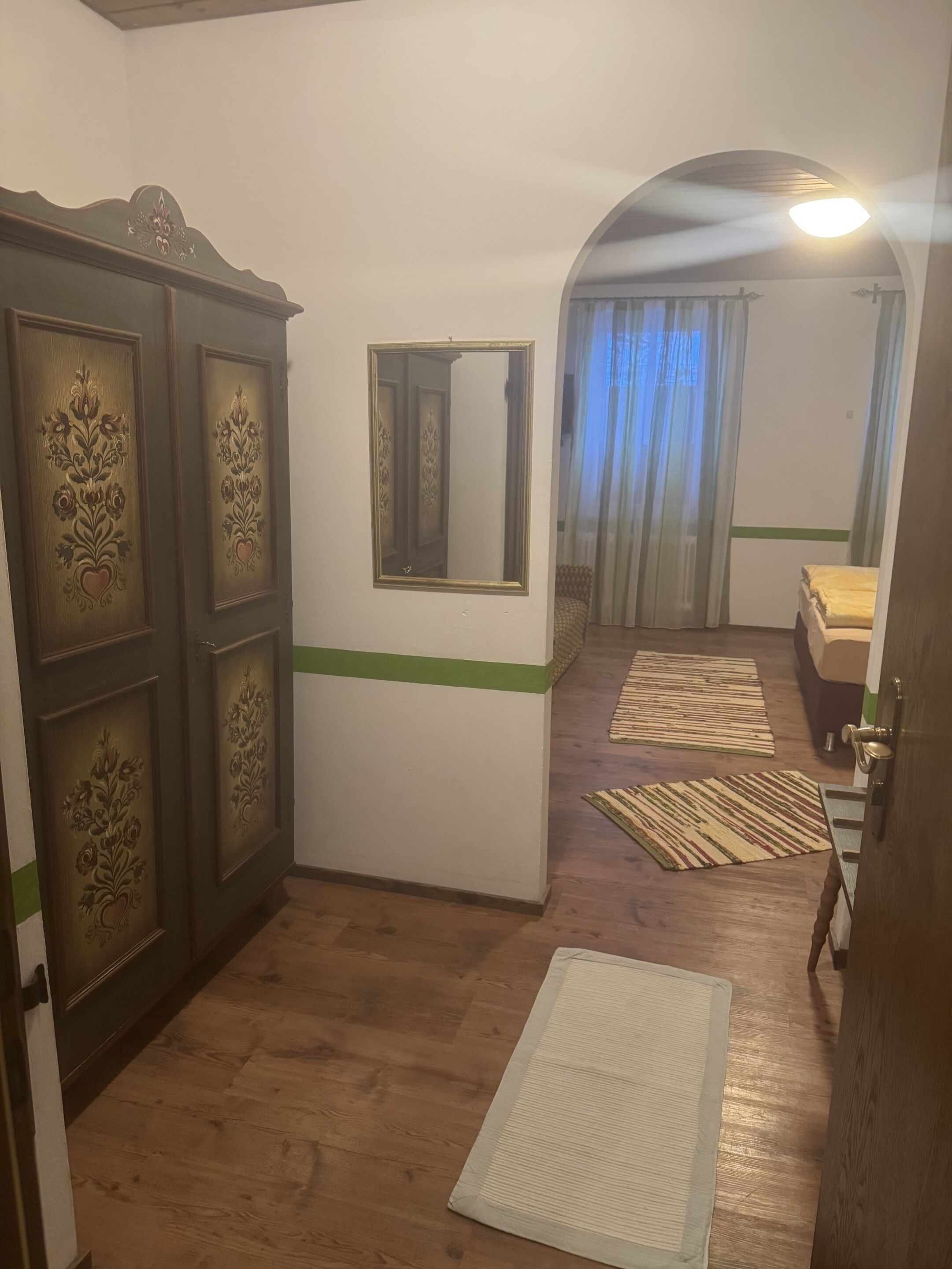 A room with a wooden wardrobe, a mirror, a bed, and a door leading to another room.